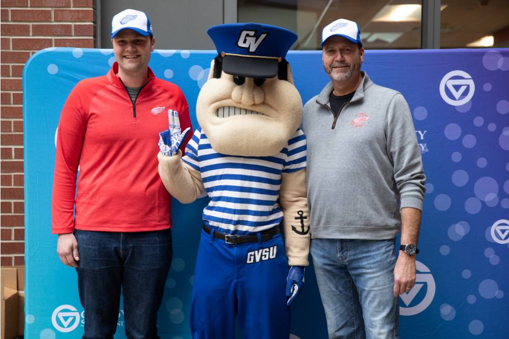 Two alumni, wearing the Red Wings GVSU hat, pose with Louie the Laker at the Detroit Red Wings GVSU Night